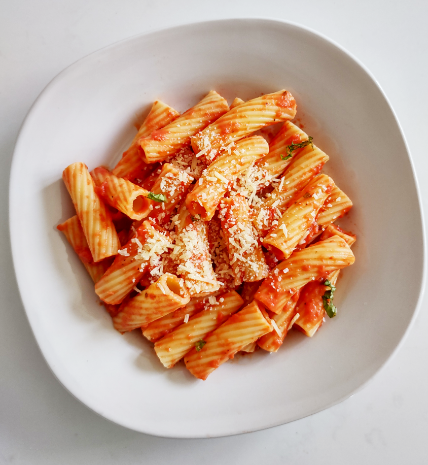 One white ceramic plate of tortiglioni pasta covered in vodka sauce, bits of herbs, and shredded parmesan cheese