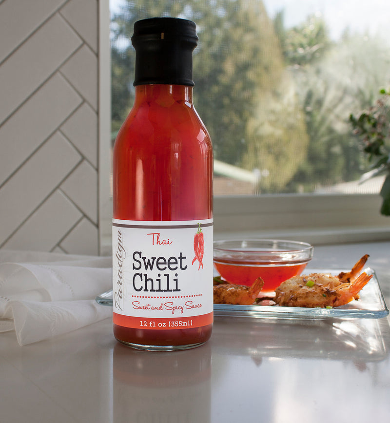 Bottle of Thai Sweet Chili on countertop in front of glass plate on which there are a couple of seasoned shrimp and a small glass bowl of thai sweet chili. Loosely rolled wax paper lays behind the plate. A window displaying trees through it is in the background.