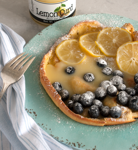 Baked Dutch baby covered in lemon curd, round lemon slices, blueberries, and powdered sugar on a light teal blue plate. A fork lays on the edge of the plate with a striped white and blue cloth. A jar of Paradigm Lemon Curd sits next to the plate.