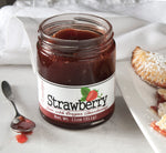 Unlidded jar of strawberry jam with jam-filled spoon to the left. To the right is a plate of strawberry hand pies dusted with powdered sugar on a white plate. Behind the jar is a crumpled white cloth.