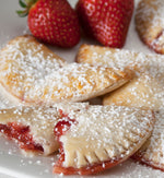 A few strawberry hand pies dusted with powdered sugar in front of two strawberries, all on a white ceramic plate.