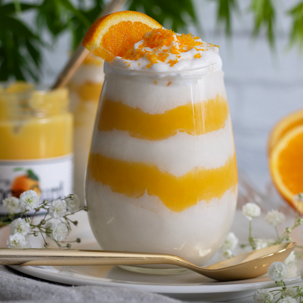 Ornate glass with layered whipped cottage cheese and Paradigm Orange Curd dessert and orange slices on a white plate with a gold spoon.