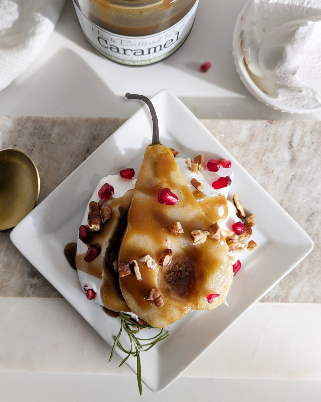 Dessert dish with poached pears, caramel sauce, and pomegranate seeds on a marble surface.
