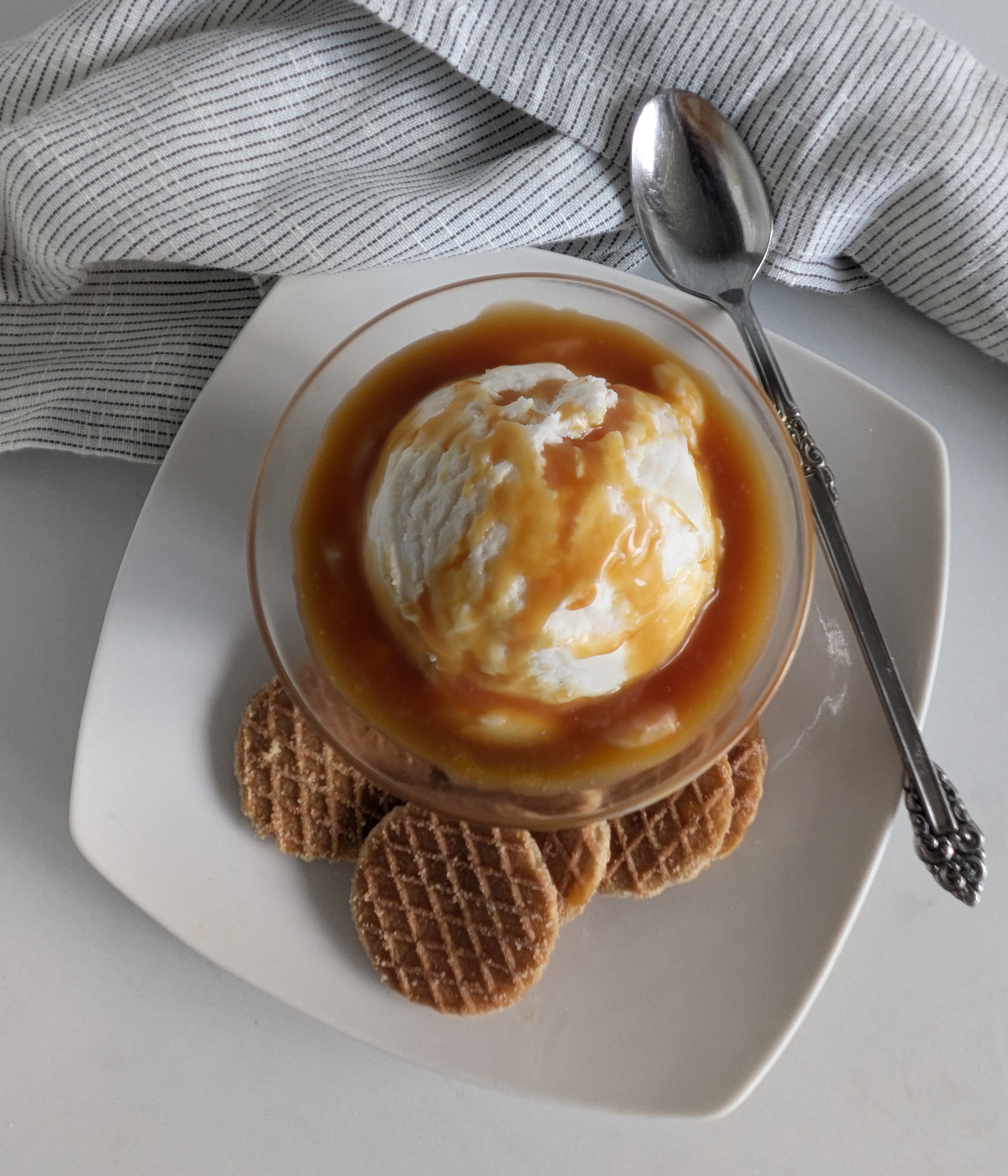 Ice cream with Paradigm Foodworks Sea Salt Caramel sauce served in a glass bowl on a white plate with waffle cookies and a spoon.