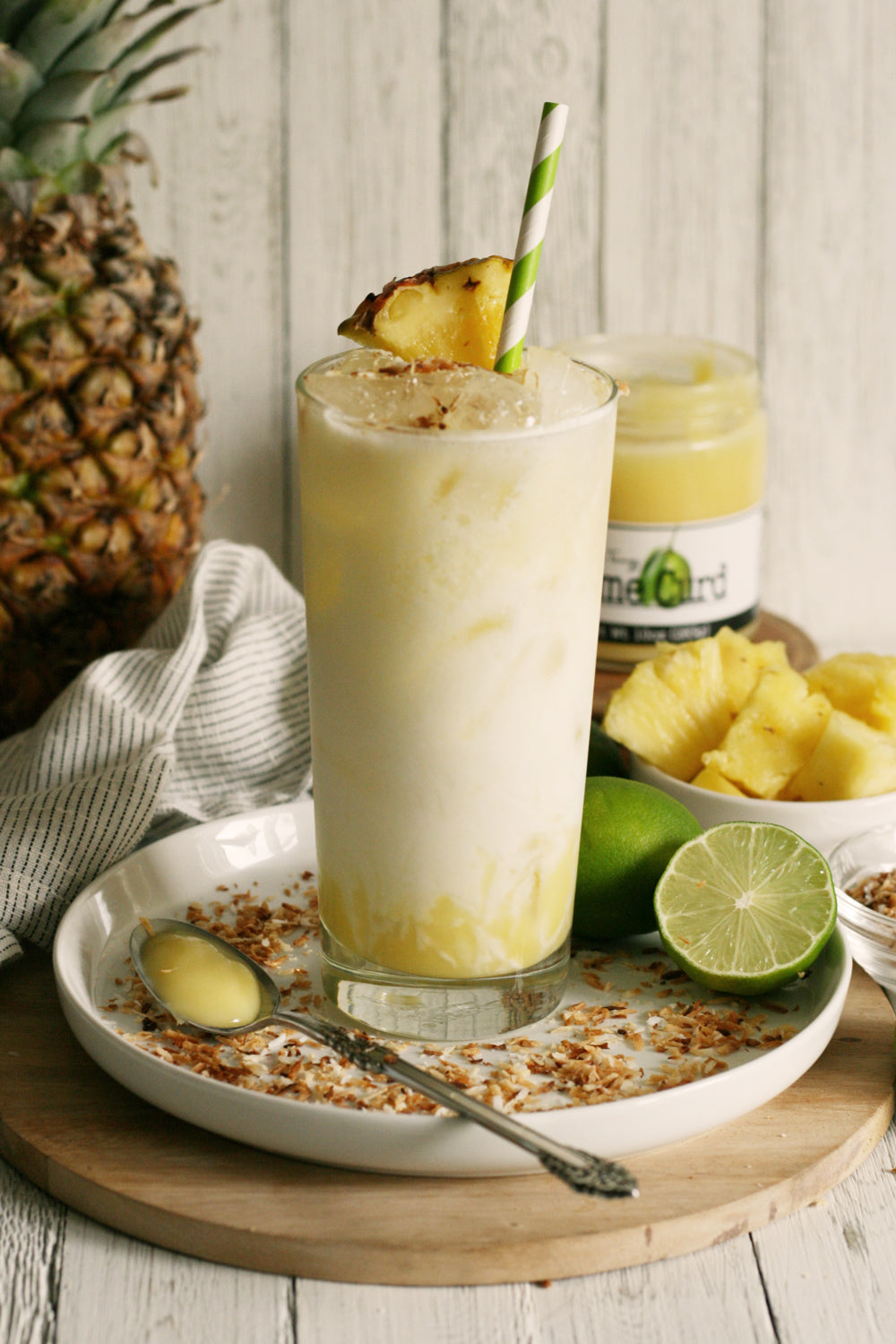 Lime Curd Pineapple smoothie with a straw on a wooden tray with a pineapple and lime in the background.