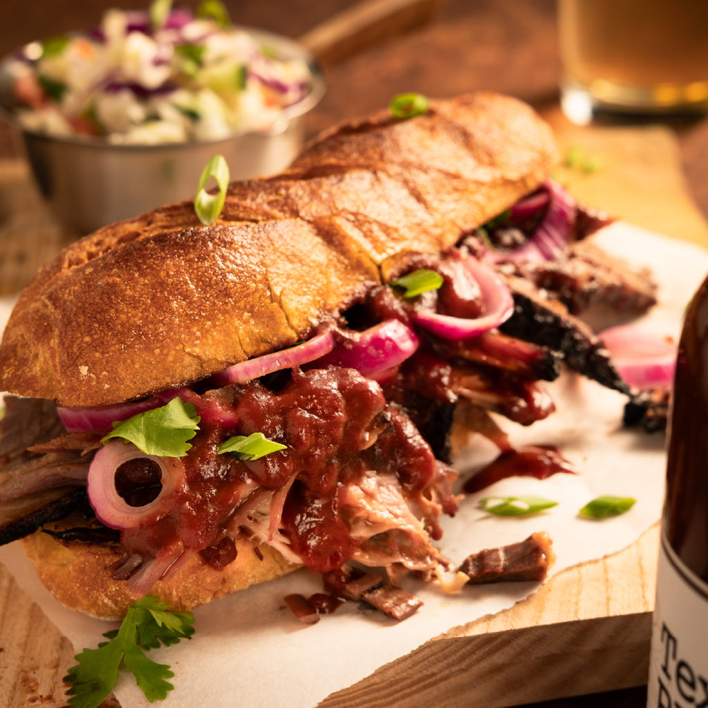 Large sandwich with pulled meat, onions, and greens on a wooden board with a side salad and glass in the background.