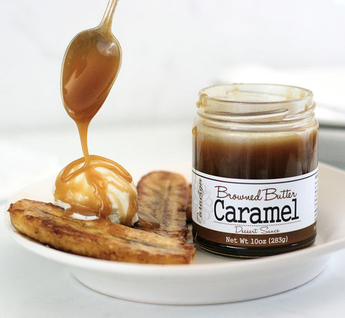Unlidded Browned Butter Caramel Sauce jar on a white plate next to fried bananas and a scoop of vanilla ice cream with a spoon above the ice cream, drizzling Browned Butter Caramel over it