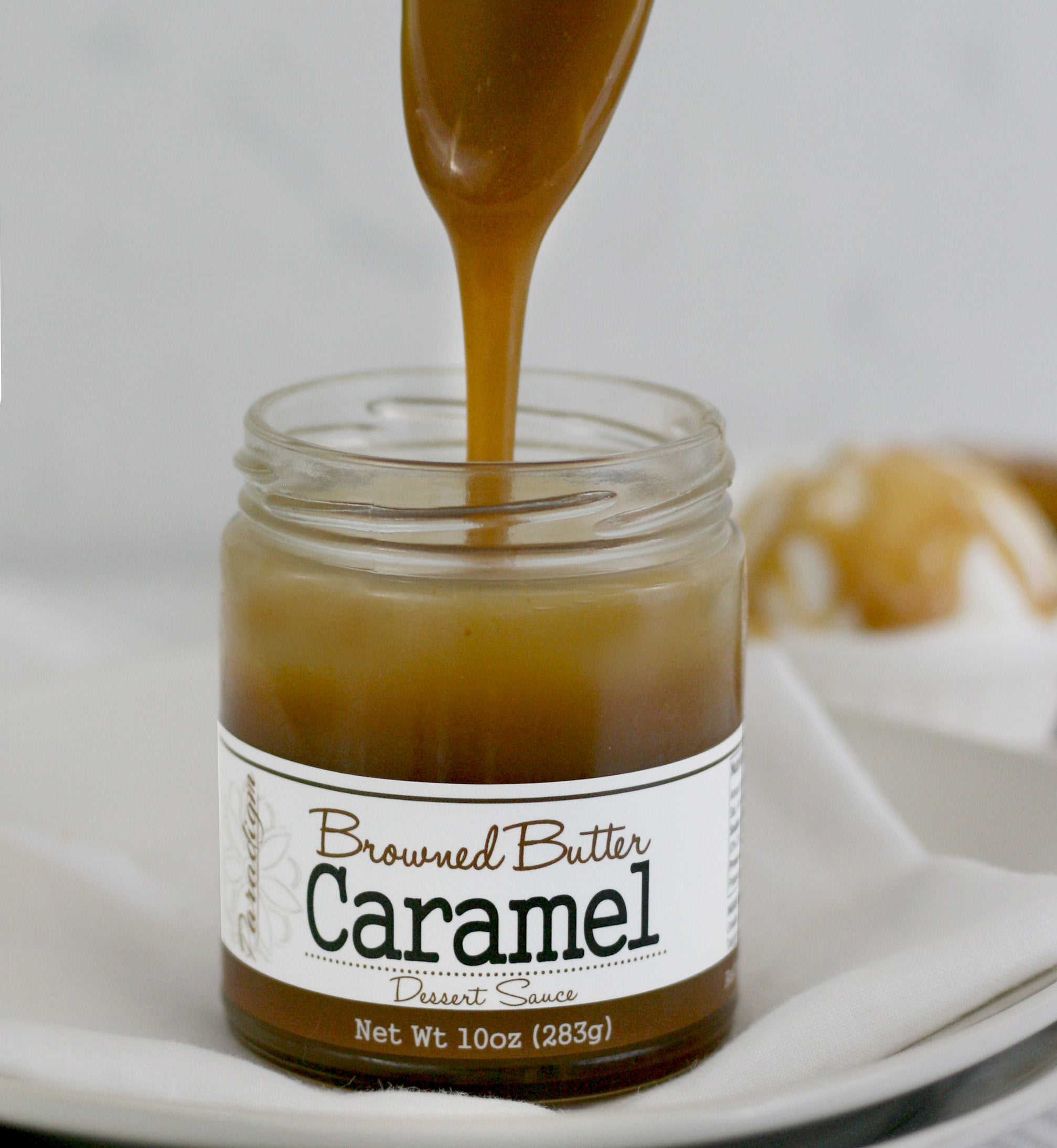 Unlidded Browned Butter Caramel Sauce jar on a linen napkin on a white plate with a spoon above the jar, drizzling Browned Butter Caramel back into it