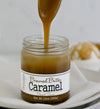 Unlidded Browned Butter Caramel Sauce jar on a linen napkin on a white plate with a spoon above the jar, drizzling Browned Butter Caramel back into it