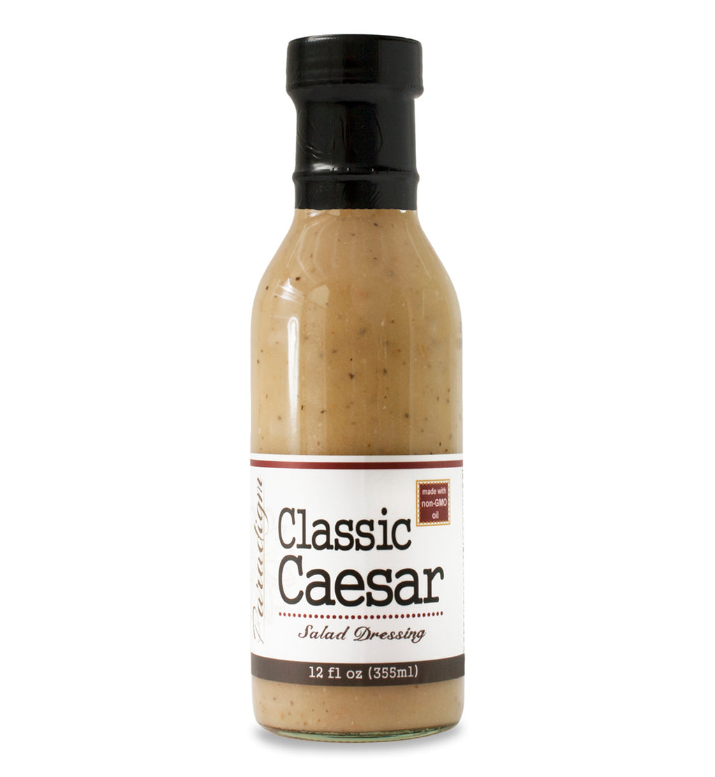 Glass ring neck bottle filled with Caesar dressing on white background. The bottle is labeled, “Paradigm Classic Caesar Salad Dressing – 12 fl oz (355ml)” and “Made with non-GMO oil”.