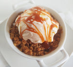 Baked granola dessert topped with vanilla ice cream with classic caramel sauce drizzled over top. The dessert is in a small ceramic bowl with handles on each side on top of a white plate