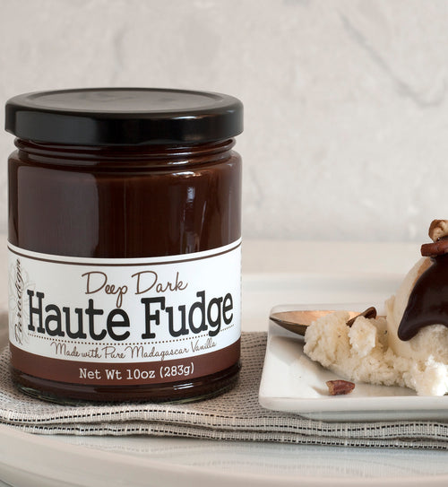Jar of Deep Dark Haute Fudge on black and white linen napkin next to a white plate. On the white plate is a scoop of vanilla ice cream topped with deep dark haute fudge and chopped nuts. A spoon lays on the plate behind the scoop of ice cream.