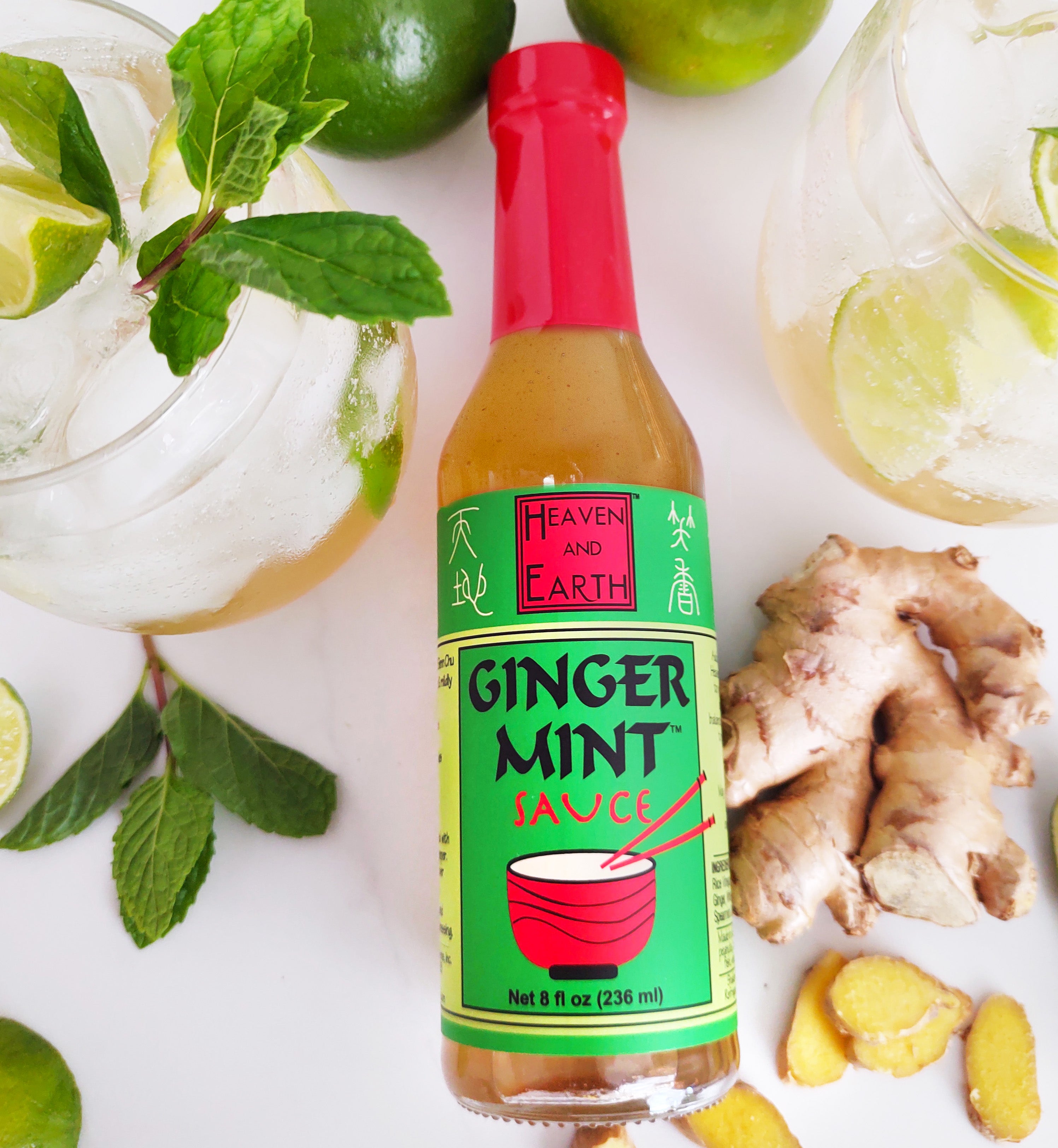 Bottle of Ginger Mint Sauce laid on white countertop, surrounded by ginger root, two ginger mint cocktails in short glasses, three limes, and a few mint leaves