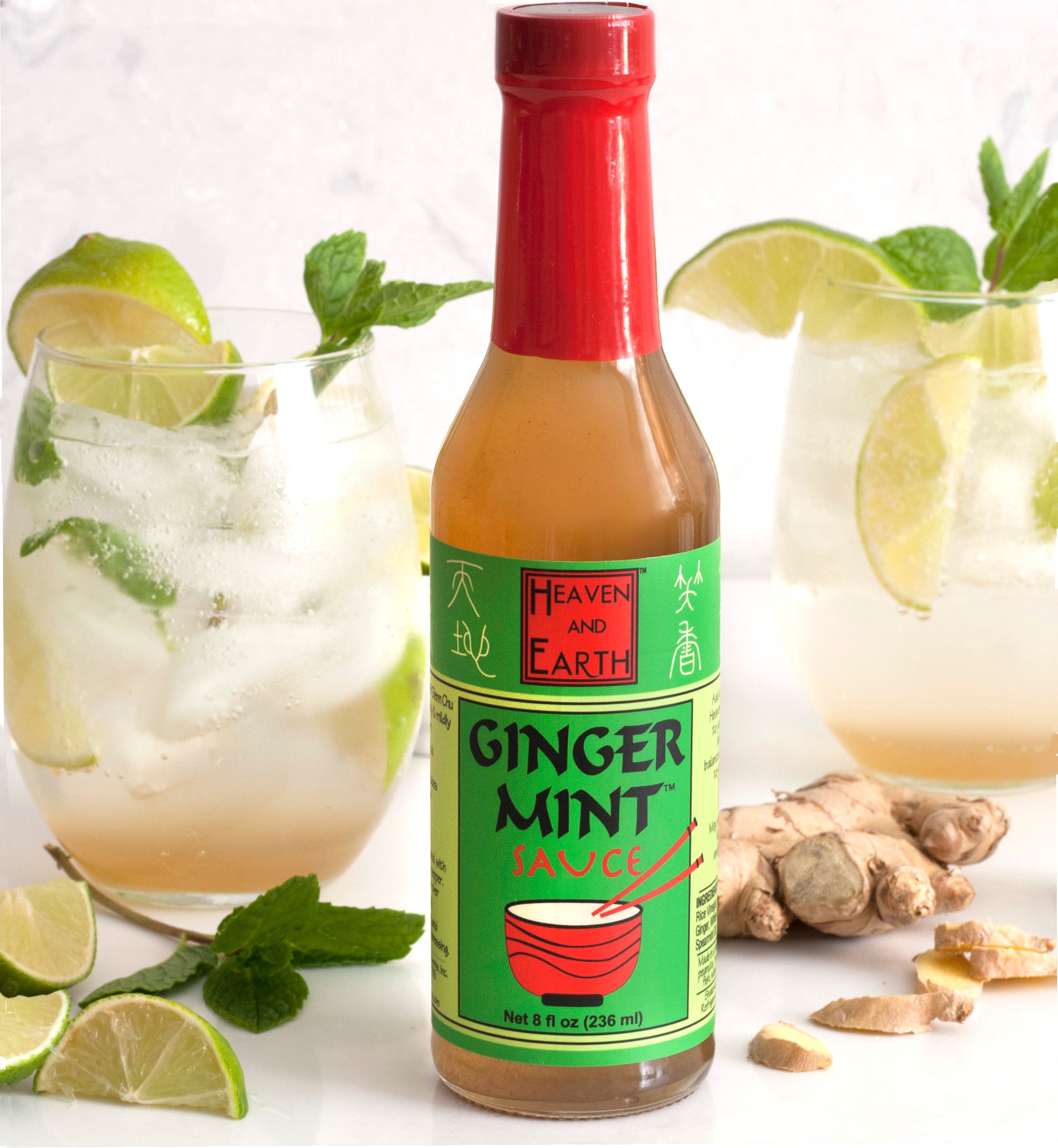 Bottle of Ginger Mint Sauce on a white countertop surrounded by two ginger mint cocktails, a few mint leaves and slices of lime, and a partially chopped ginger root.