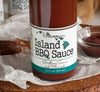 Bottom half of bottle full of Hawaiian Island BBQ sauce on crinkled wax paper on top of wood round. Behind the bottle is a glass bowl of BBQ sauce, a couple more bottles of barbecue sauce, and a basting brush covered in BBQ sauce.