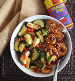 Overhead view of a bowl of shrimp and vegetables covered in Dragon Fire Chili Sauce and Sesame seeds, with a spoon tucked in the side. The bowl sits on a brown woven table with a crumpled burlap cloth and a bottle of dragon fire chili sauce. 