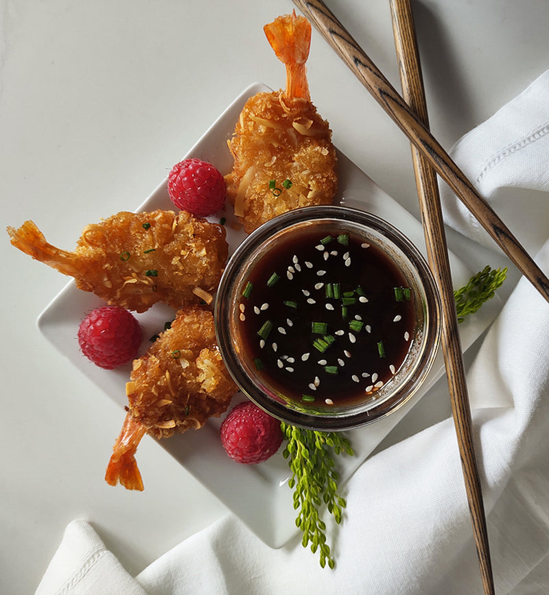 Bowl of raspberry hoisin sauce topped with sesame seeds and chopped chives in the center of a white square plate, surrounded by deep-fried shrimp, a few raspberries, and a few sprigs of greenery. A set of chopsticks lean crisscrossed on one side of the plate with a white cloth draped underneath them.