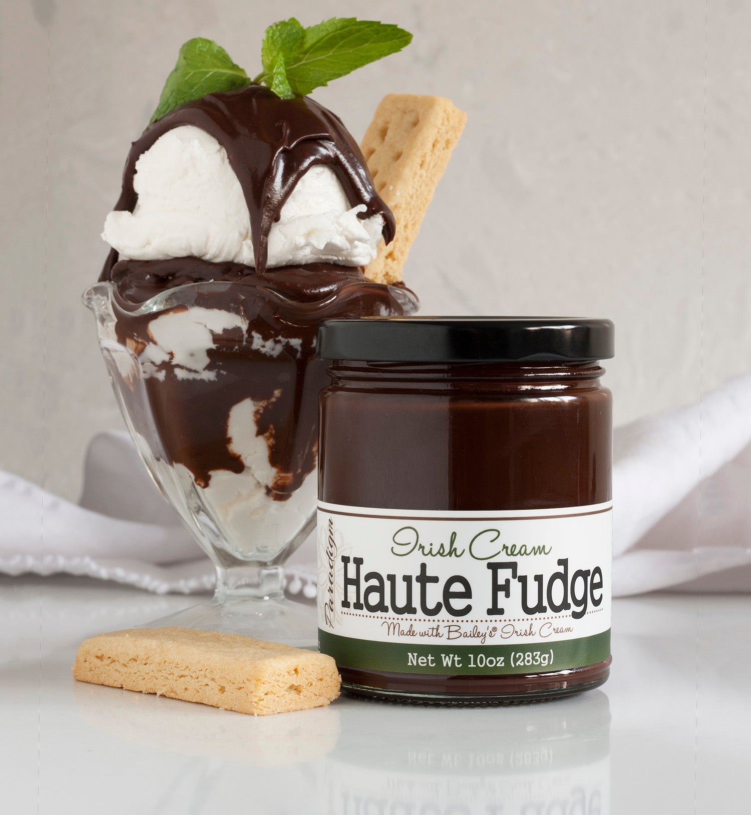 Irish Cream Haute Fudge on reflective countertop with biscuit cookie next to it. Behind it is a hot fudge sundae using Irish Cream Haute Fudge, topped with mint leaves and another long biscuit cookie in the side of the sundae. A white cloth is laid behind the sundae. 