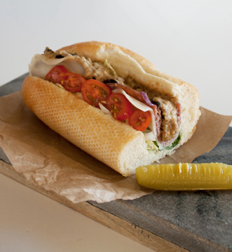 Half a sub sandwich on a brown napkin on top of a piece of wood. The sandwich contains tomatoes, meat, sauce, and more, and next to it there is a quarter pickle cut lengthwise.