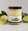 Jar of Paradigm Lime Curd on white countertop with dark green leaves and lime slices on either side. Background is a white marble wall.