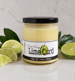 Jar of Paradigm Lime Curd on white countertop with dark green leaves and lime slices on either side. Background is a white marble wall.