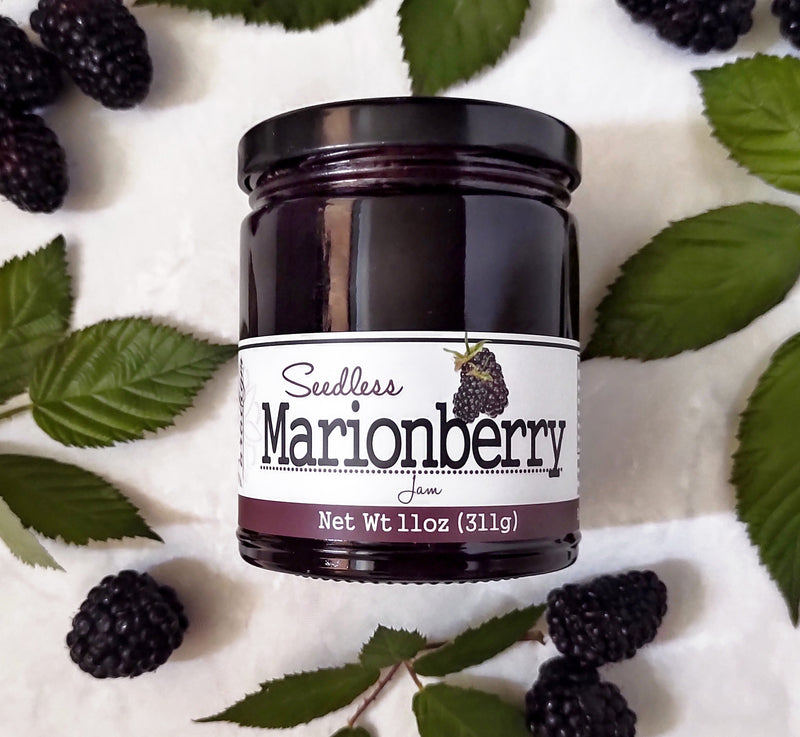 Jar of Seedless Marionberry Jam laying on white countertop surrounded by marionberries and marionberry leaves.