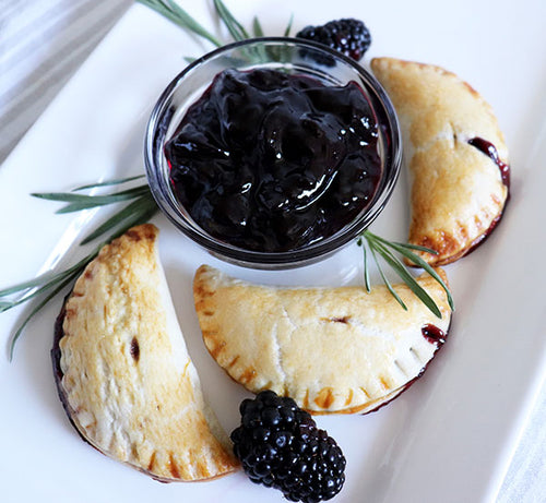 Glass bowl filled with marionberry jam next to marionberry hand pies, sprigs of rosemary, and two marionberries. All of these sit on a white rectangular plate.