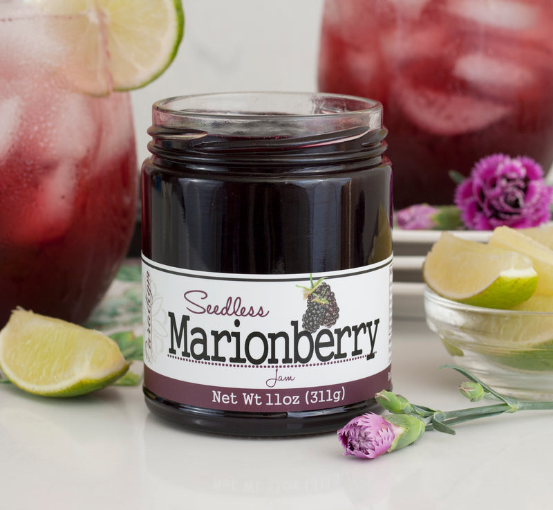 Unlidded jar of marionberry jam with purple carnation in front of jar. Two glass pitchers of marionberry lemonade sit behind jar, with slices of lime in and around the pitchers. Next to the jar is a small bowl of lime slices. 
