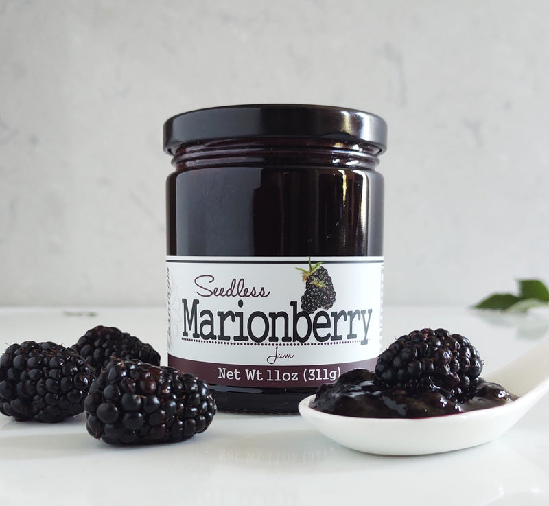 Jar of Seedless Marionberry Jam in front of ceramic spoon filled with jam and a single marionberry. Three loose marionberries sit on the counter to the left of the jar.