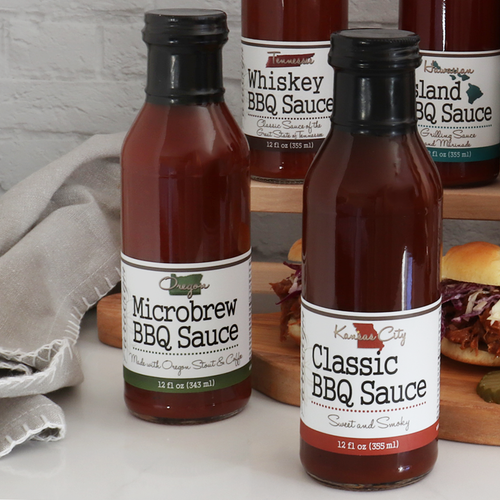 Bottom half of bottle full of Oregon Microbrew BBQ Sauce on crinkled wax paper on top of wood round. Behind the bottle is a glass bowl of BBQ sauce, a couple more bottles of barbecue sauce, and a basting brush covered in BBQ sauce.