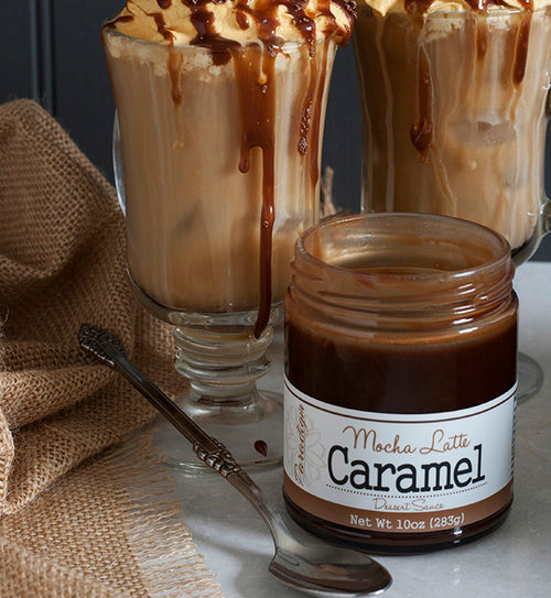 Two glass mugs filled with decadent iced mochas topped with whipped cream and mocha latte caramel sauce drizzled over top and running down the sides of the mugs. A single spoon and a Mocha Latte Caramel Sauce jar are in front of the mochas, and a crumpled burlap cloth is beside them