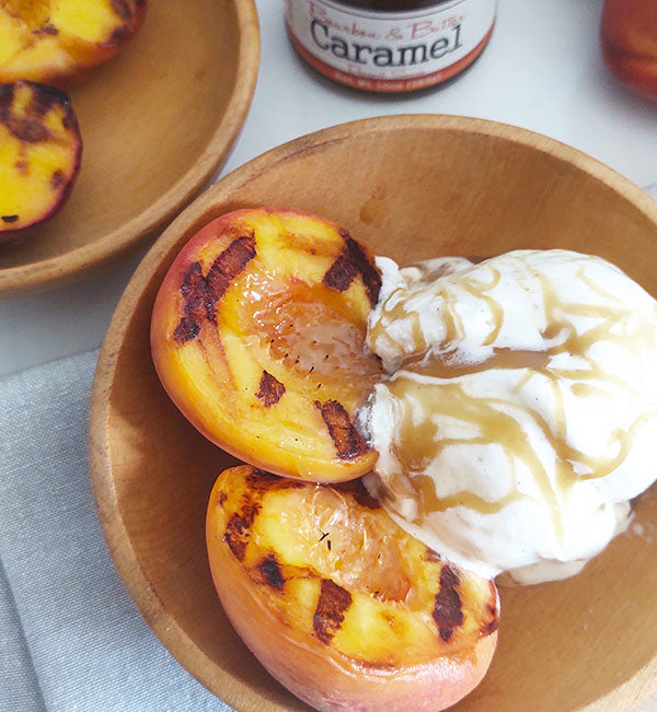 Wooden bowls on grey linen napkins with jar of Bourbon & Butter Caramel next to them. The wooden bowls contain grill-seared peaches and vanilla ice cream, with Bourbon & Butter Caramel drizzled over them.