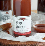 Bottom half of bottle full of NC Mop Sauce on crinkled wax paper on top of wood round. Behind the bottle is a glass bowl of mop sauce, a couple more bottles of barbecue sauce, and a basting brush covered in mop sauce.