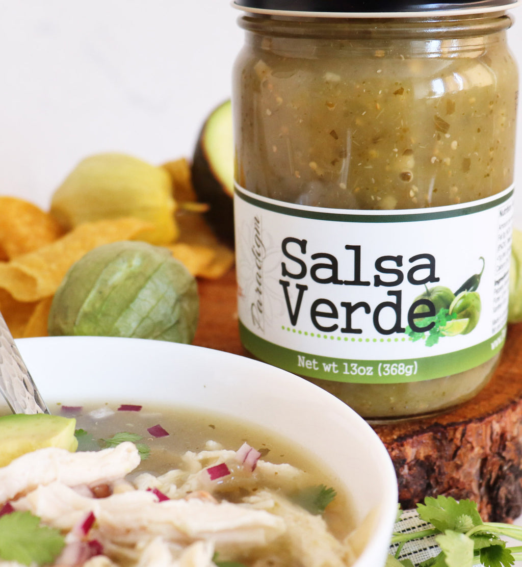 Jar of salsa verde on natural wood round next to peppers and tortilla chips and half and an avocado. A white bowl of tortilla soup sits in the foreground. 