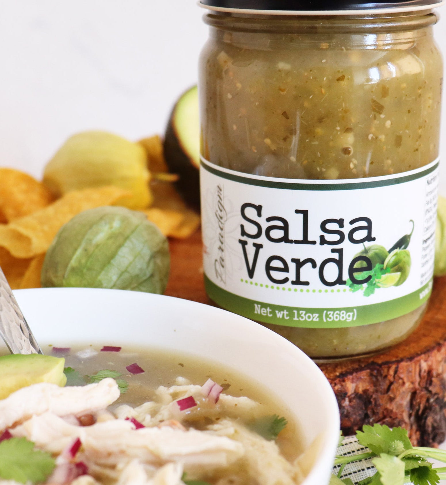 Jar of salsa verde on natural wood round next to peppers and tortilla chips and half and an avocado. A white bowl of tortilla soup sits in the foreground. 