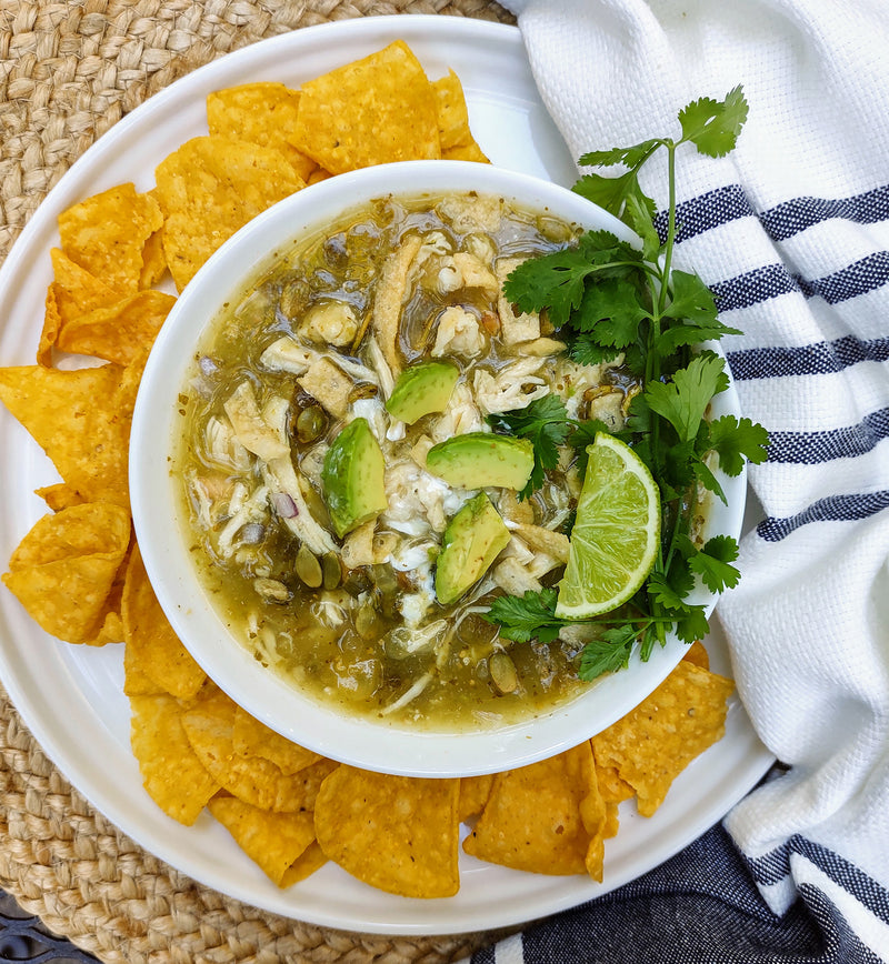 White bowl of tortilla soup topped with salsa verde, avocado, lime, and cilantro on white plate covered in tortilla chips. The plate is partially covered by a draped white and blue striped cloth and sits on a braided straw placemat. 