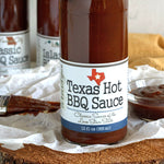 Bottom half of bottle full of Texas Hot BBQ Sauce on crinkled wax paper on top of wood round. Behind the bottle is a glass bowl of BBQ sauce, a couple more bottles of barbecue sauce, and a basting brush covered in BBQ sauce.