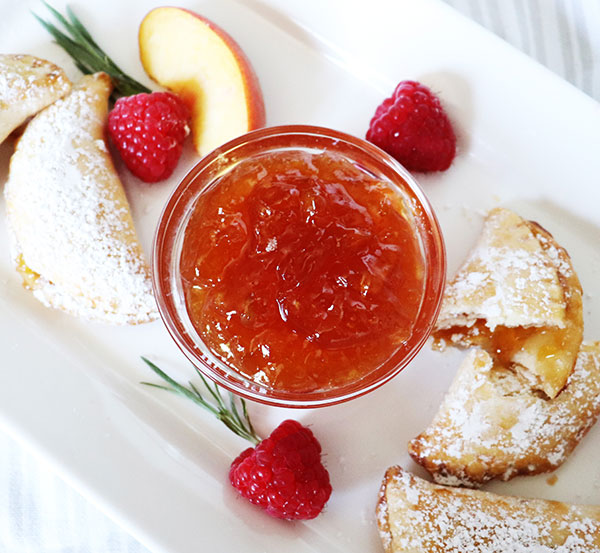 White rectangular plate on top of which is a small glass bowl of peach raspberry jam, surrounded by a few peach raspberry hand pies, bits of rosemary, and a few raspberries.