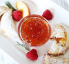 White rectangular plate on top of which is a small glass bowl of peach raspberry jam, surrounded by a few peach raspberry hand pies, bits of rosemary, and a few raspberries.