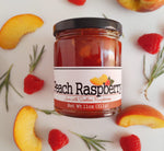 Jar of peach raspberry jam laying in the center of the image surrounded by a few peach slices, raspberries, and pieces of rosemary.