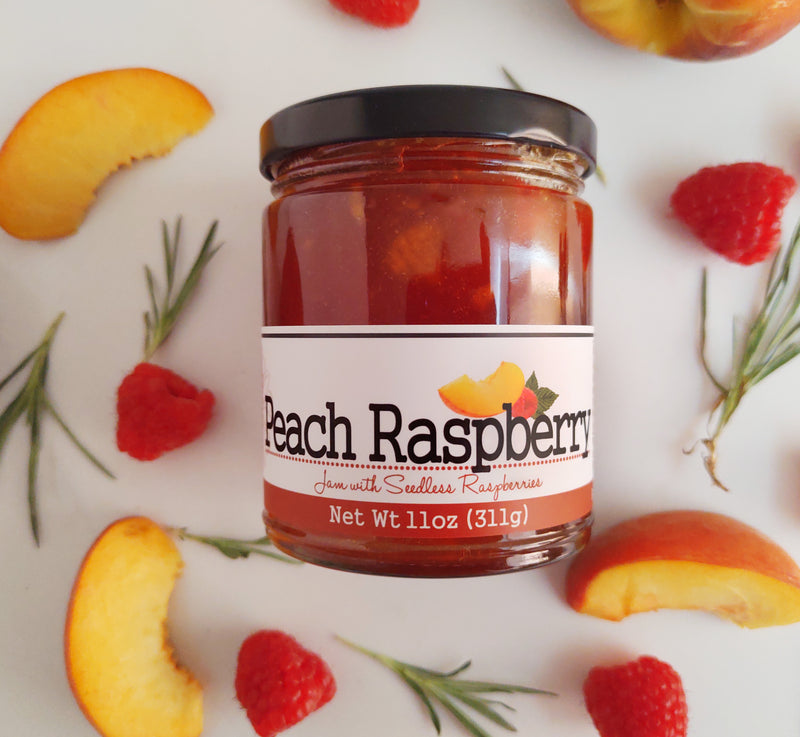 Jar of peach raspberry jam laying in the center of the image surrounded by a few peach slices, raspberries, and pieces of rosemary.