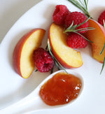 Closeup of a few peach slices on a white ceramic plate with a few pieces of rosemary and a few raspberries scattered on the plate and a white ceramic spoon containing peach raspberry jam.