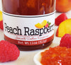 Closeup of the lower half of a jar of peach raspberry jam with peaches and raspberries strewn about around it and a ceramic spoon full of the jam in the foreground.