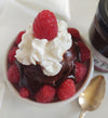 Closeup overhead view of hot fudge sundae in a white cup. The cup contains a scoop of ice cream surrounded by a circle of raspberries, and is covered in Raspberry Haute Fudge.  The fudge is topped with a dollop of whipped cream and one raspberry. Beside the sundae is a jar of Raspberry Haute Fudge and a spoon.