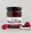 Jar of raspberry Haute Fudge on a countertop with a single raspberry on the left side and a serving spoon filled with Raspberry Haute Fudge and a raspberry on the right side