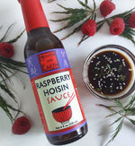 Bottle of Raspberry Hoisin Sauce laying on white countertop next to small glass bowl of raspberry hoisin sauce topped with sesame seeds and greens. The bowl and bottle are surrounded by scattered Japanese maple leaves and raspberries. 