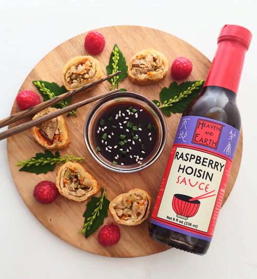 Bowl of raspberry hoisin sauce topped with sesame seeds and chopped chives in the center of round wooden cutting board. Round Slices of spring rolls surround the bowl, along with a few raspberries, leaves, sprigs of greenery, a set of chopsticks, and a bottle of Raspberry Hoisin Sauce. 