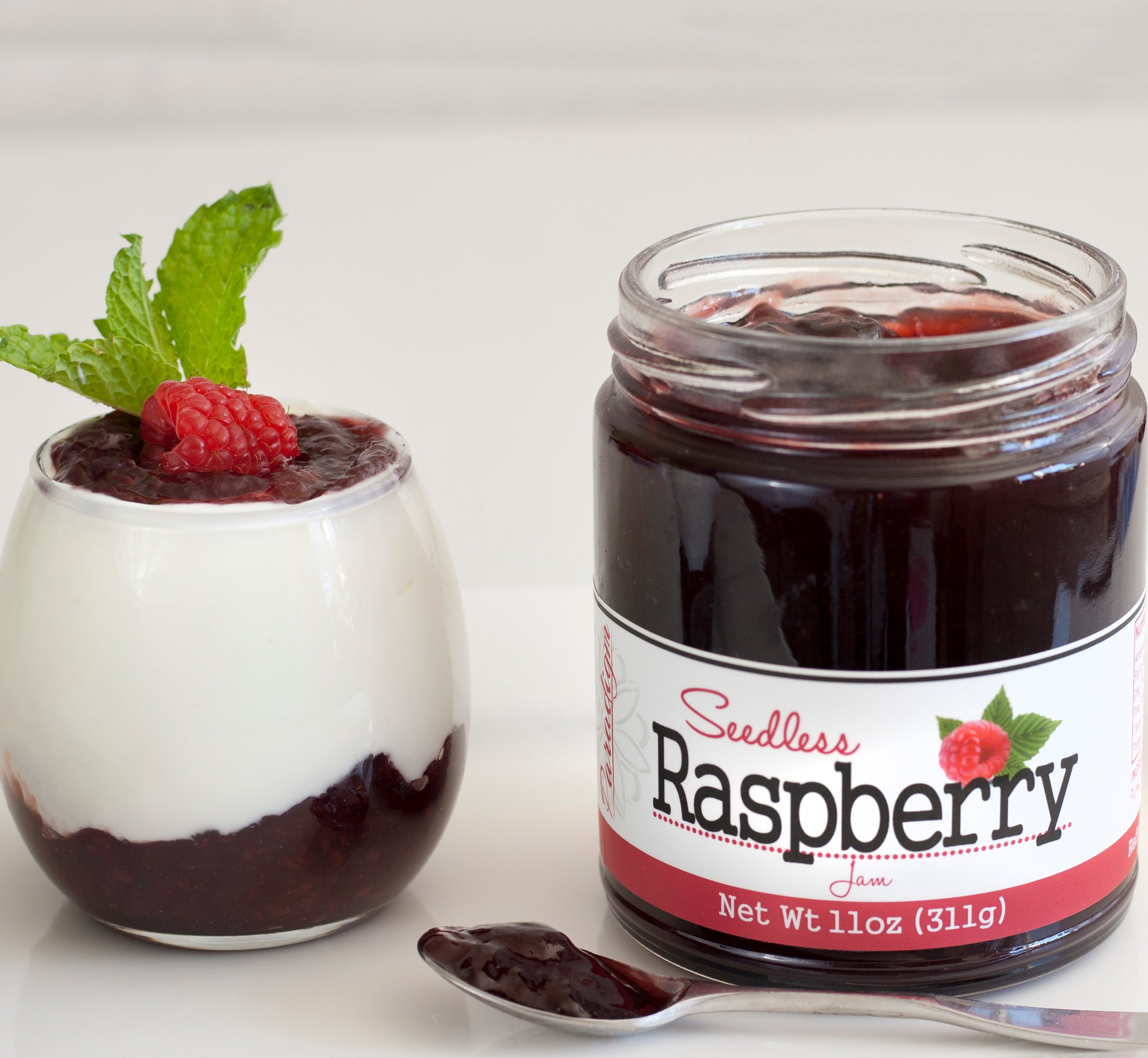 Unlidded jar of Seedless Raspberry Jam on white countertop next to glass cup of vanilla yogurt and jam, topped with mint leaves and one raspberry. In front of the jar is a spoon full of jam.