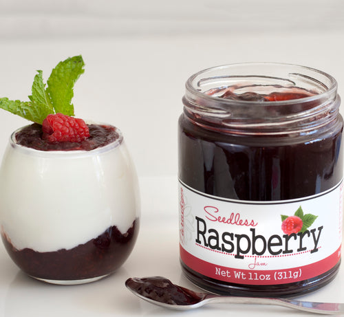 Unlidded jar of Seedless Raspberry Jam on white countertop next to glass cup of vanilla yogurt and jam, topped with mint leaves and one raspberry. In front of the jar is a spoon full of jam.
