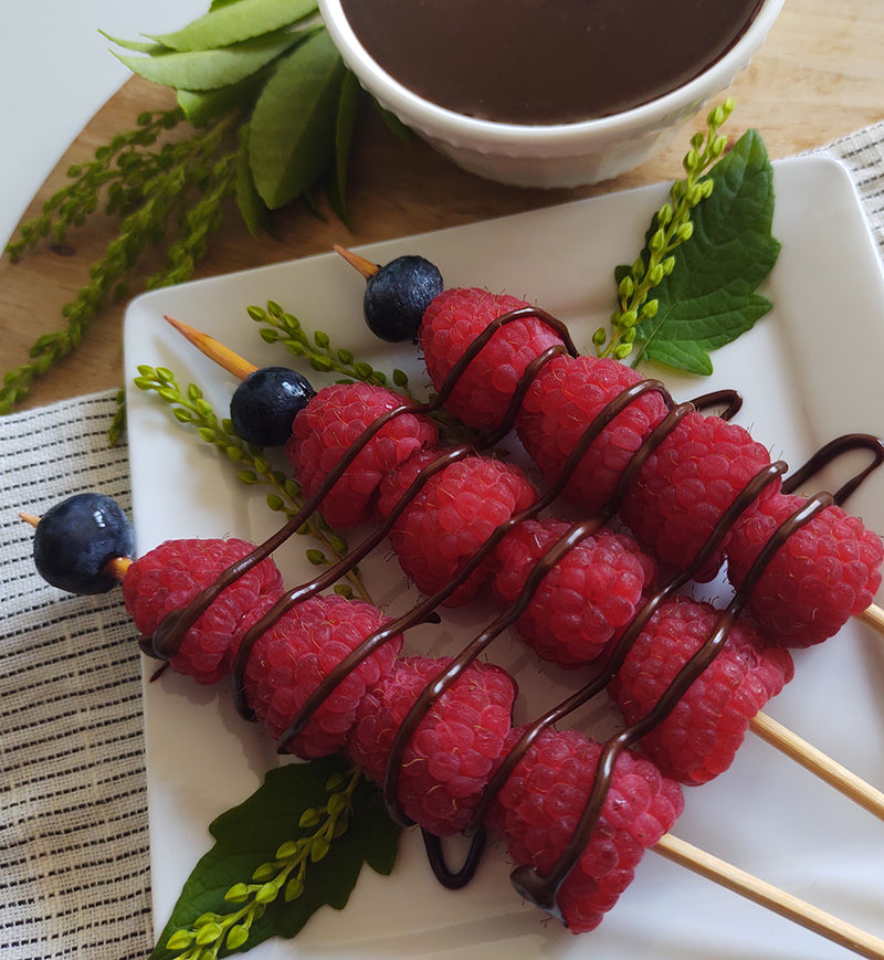White square plate with greenery and three fruit skewers on it. The skewers are mostly raspberries with one blueberry each and are drizzled with Raspberry Haute Fudge. Next to the plate of skewers is a folded black and white linen napkin, more greenery, and a white ceramic bowl of Raspberry Haute Fudge. 